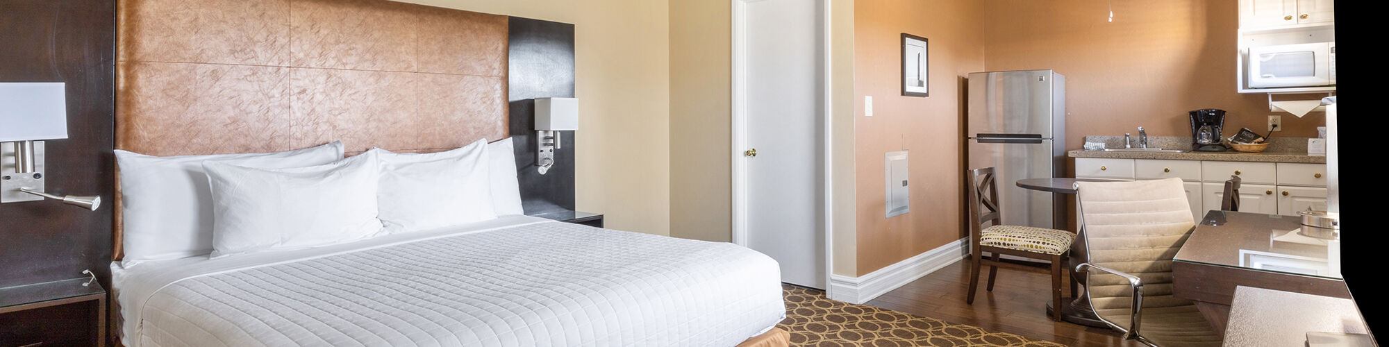 The image shows a hotel room with a bed, a desk, a chair, a TV, and a kitchenette with a microwave and coffee maker on a patterned carpet.
