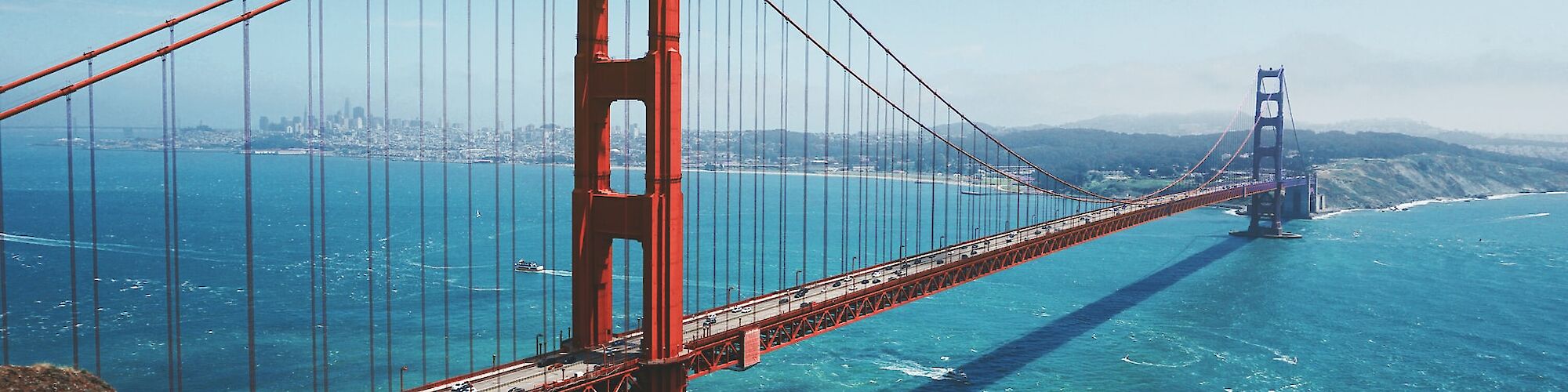A famous red suspension bridge spans across a blue body of water, with a distant city skyline visible under a hazy sky in the background.