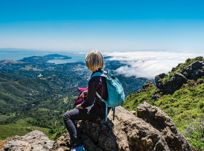 A person with a blue backpack sits on a rocky ledge, overlooking a scenic view of mountains, hills, and a coastline under a clear blue sky.