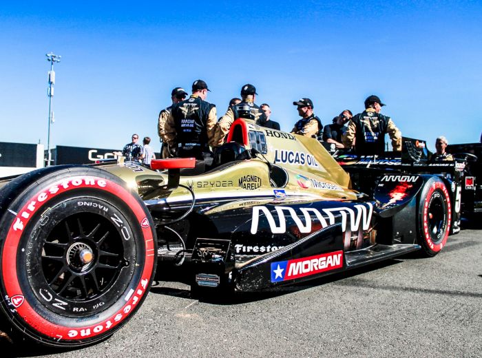An IndyCar race car with "Arrow" branding is on the track, surrounded by team members in racing uniforms under a clear blue sky.