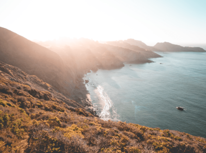 This image portrays a serene coastal scene with rugged cliffs meeting a calm ocean under a bright, clear sky, bathed in warm sunlight.