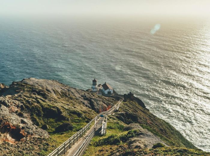 A lighthouse located on a rugged cliff by the ocean with a pathway leading to it. The scene captures a clear, sunny day with calm waves.
