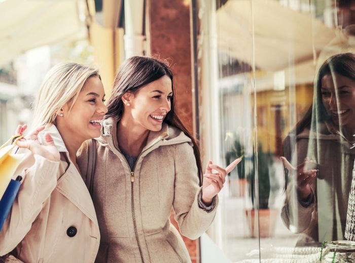 Two women smiling and holding shopping bags, looking at a store window display, appearing thrilled and engaged in conversation.