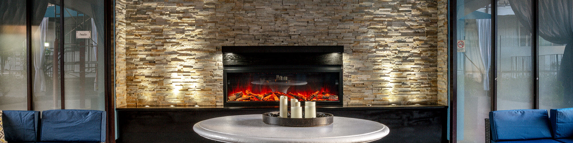 A cozy lounge area with wicker furniture, a round table, pendant lights, and a modern fireplace against a brick wall, providing a warm ambiance.