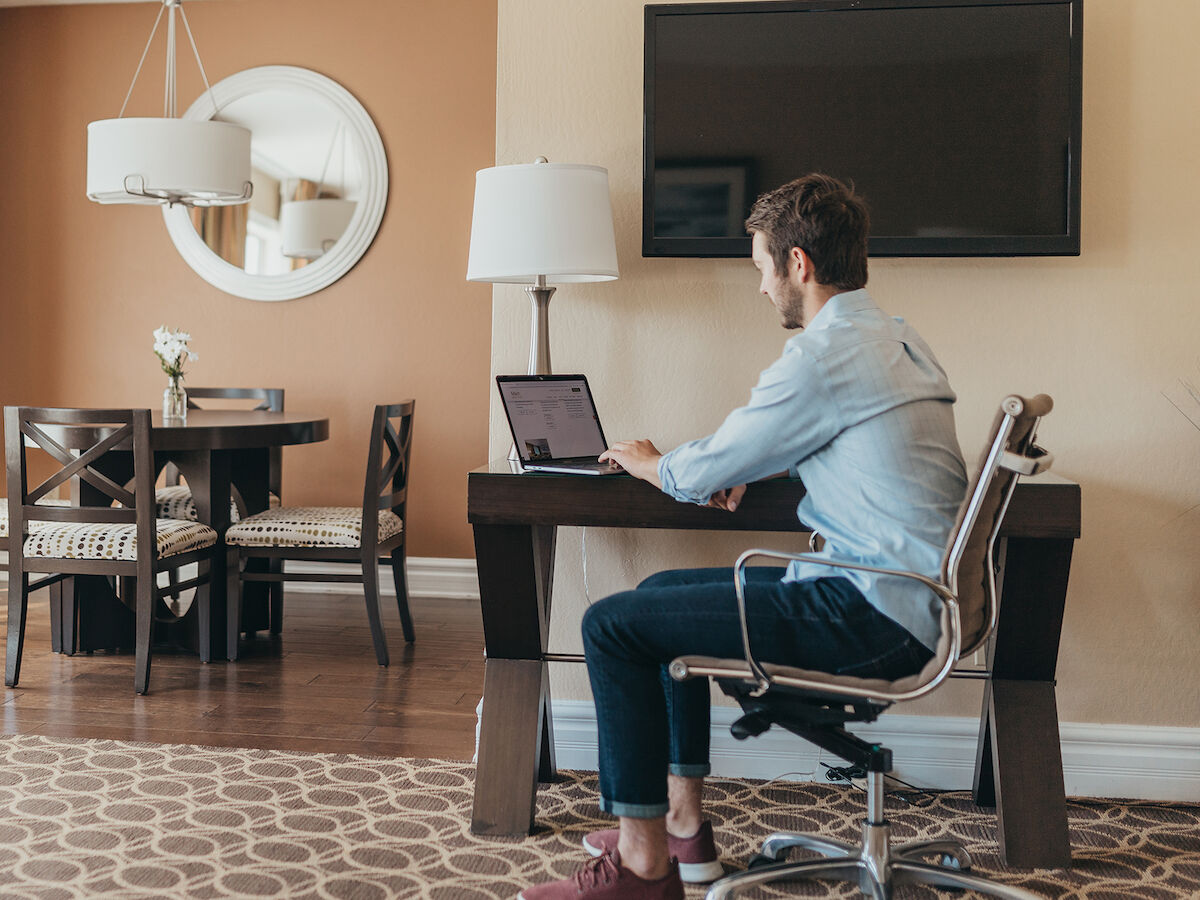 A person is sitting at a desk working on a laptop in a well-decorated room with a dining area and TV on the wall.