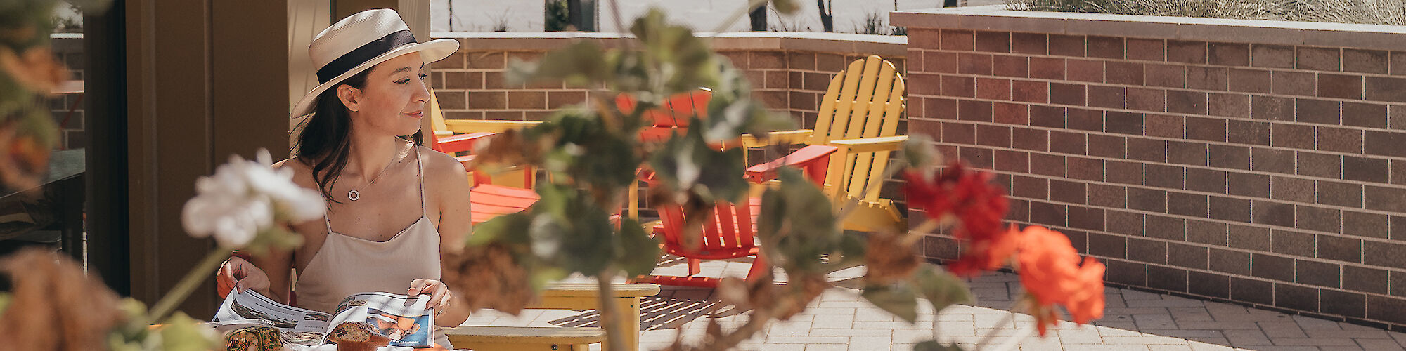 A woman wearing a hat sits at an outdoor café table with flowers in the foreground and yellow chairs in the background, enjoying a sunny day.