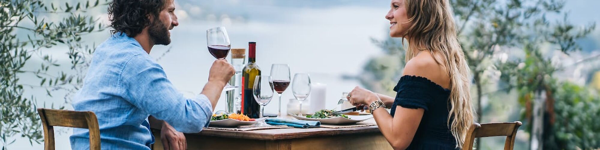 A man and woman enjoy an outdoor meal with wine, overlooking a scenic landscape of mountains and water, seated at a wooden table.