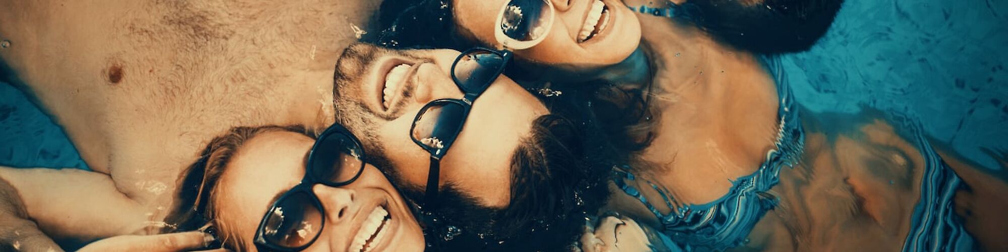 Four people wearing sunglasses are floating in a pool, smiling at the camera, creating a friendly and relaxed atmosphere.