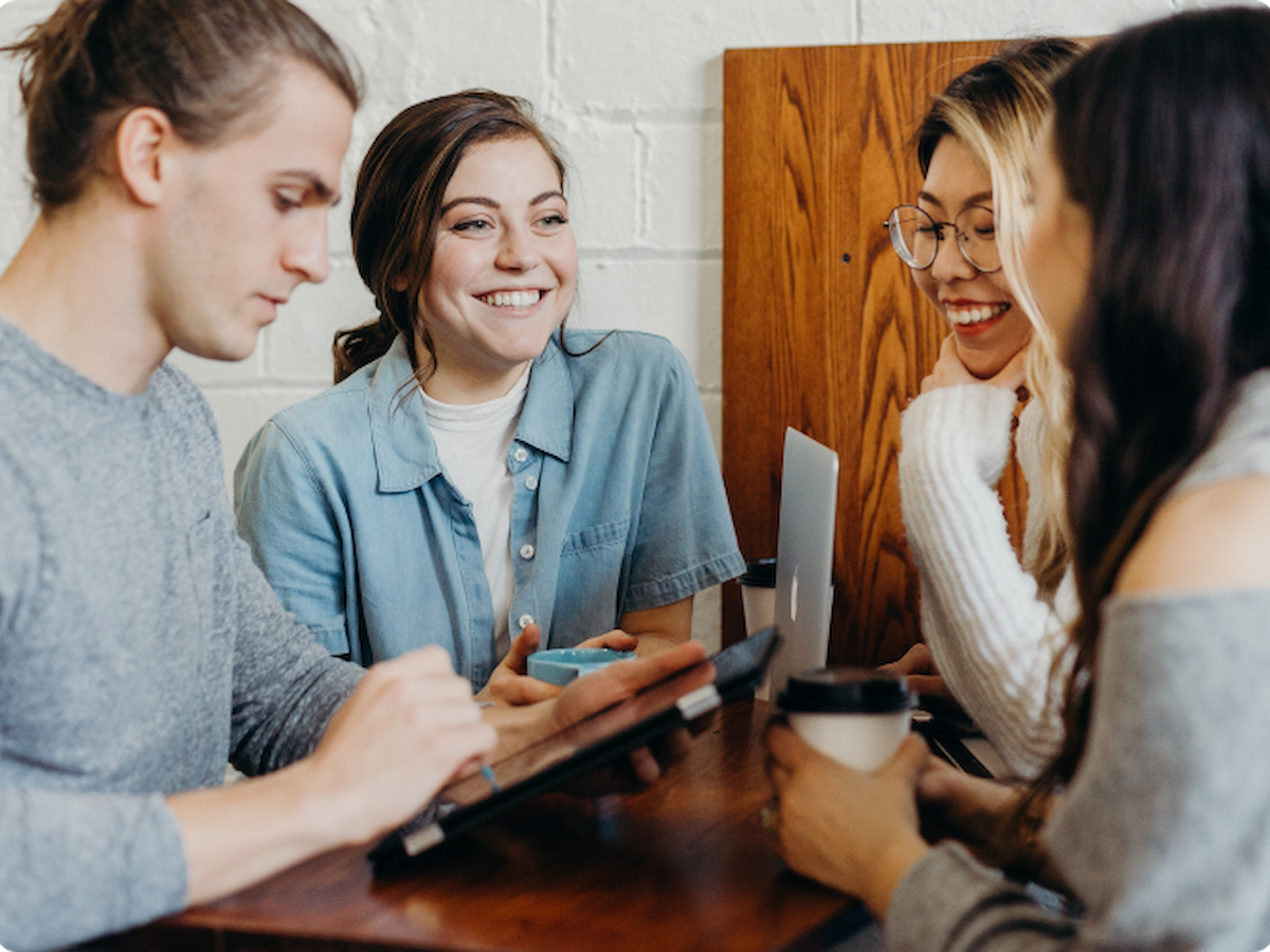 A group of four people are sitting around a table, interacting and smiling, one holding a coffee cup and another using a tablet, with a laptop visible.
