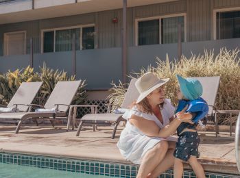 A woman and child in sun hats are sitting by the edge of a swimming pool outside a building, with lounge chairs in the background.
