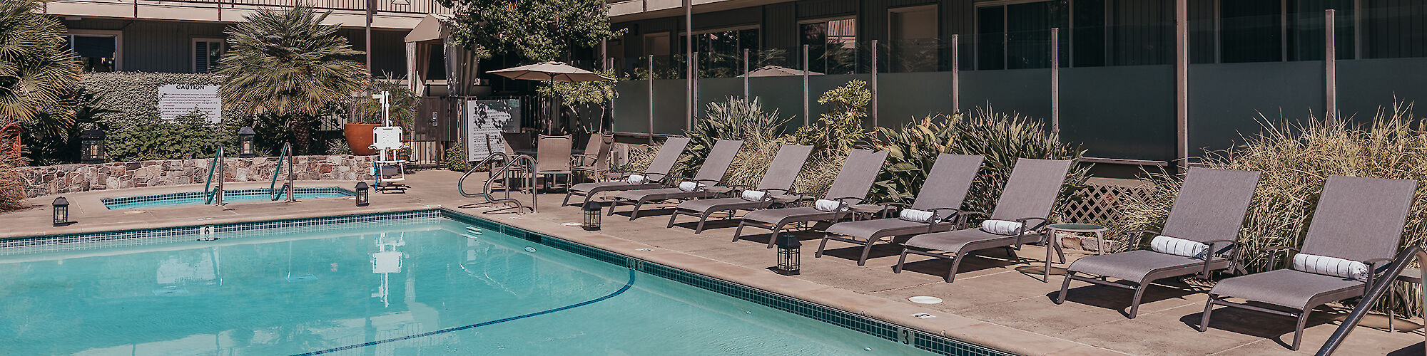 The image shows a swimming pool area with lounge chairs, a jacuzzi, and surrounding apartment buildings with balconies and greenery in the background.
