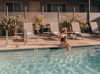 A woman in a black swimsuit and hat sits by the edge of a pool with lounge chairs and plants in the background.