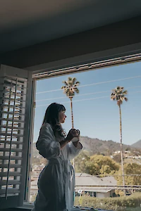 A person in a white robe is standing in front of an open window, holding a cup and gazing outside at a scenic view with mountains and palm trees.