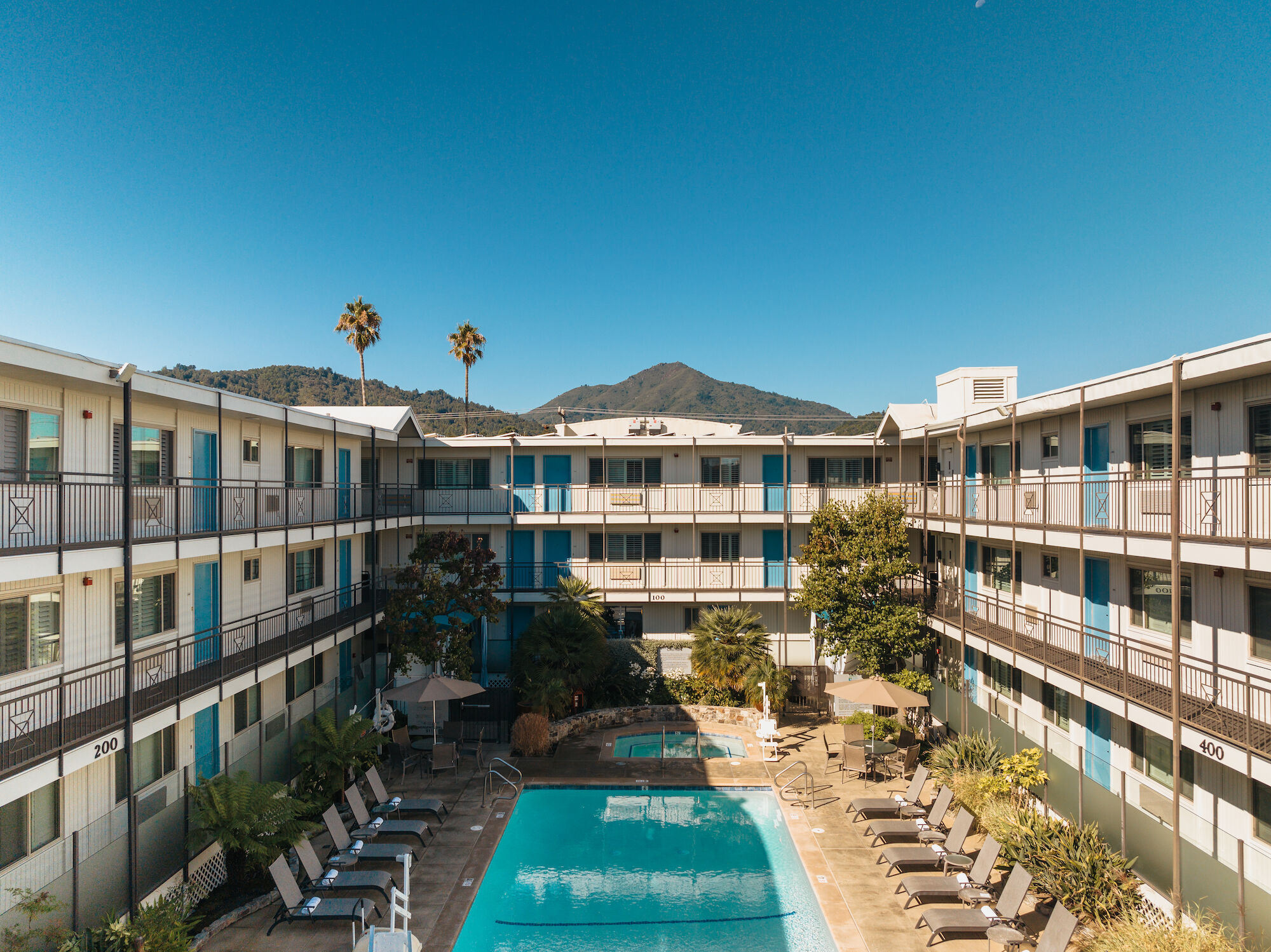 The image shows a sunny day at a courtyard-style hotel or apartment complex with a central swimming pool, surrounded by lounge chairs and outdoor seating.