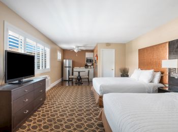 The image shows a hotel room with two beds, a dresser with a TV, a kitchenette, and patterned carpet.