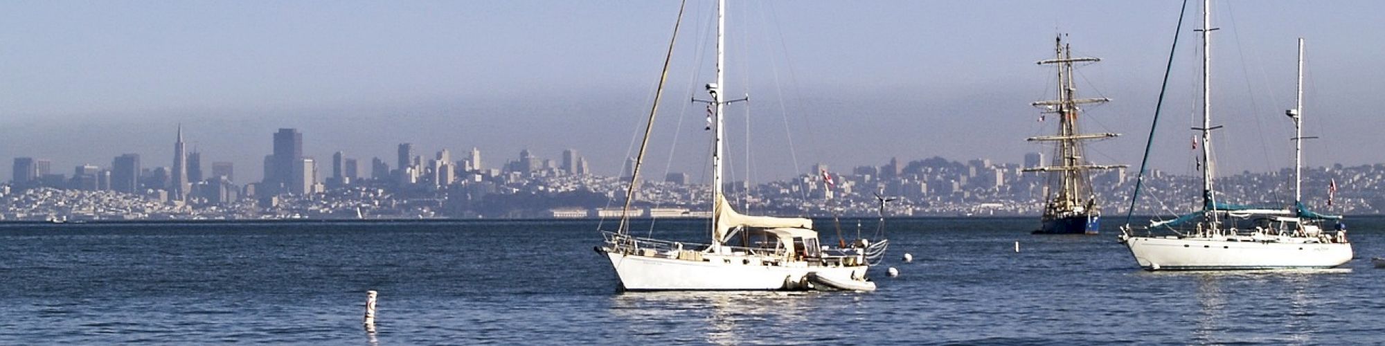 The image shows sailboats on a body of water, with a rocky shoreline and city skyline in the distance under a clear blue sky.