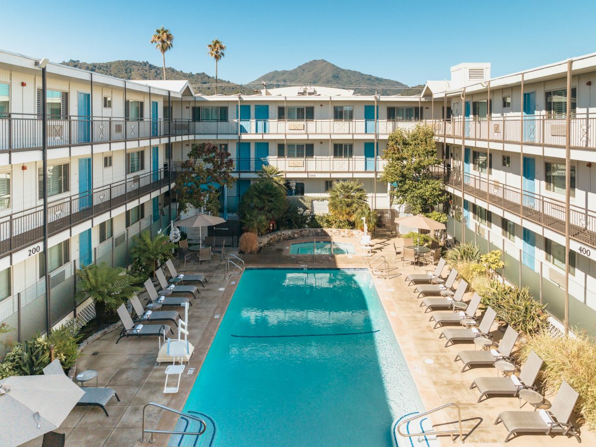 The image shows a hotel courtyard with a central swimming pool, surrounded by lounge chairs and greenery, set against mountain views.