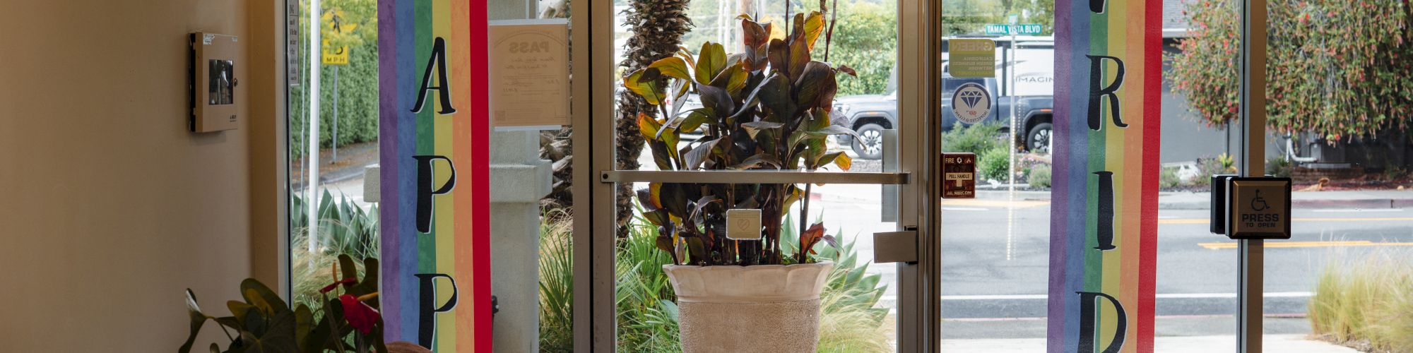 The image shows a doorway with rainbow banners spelling "HAPPY PRIDE," flanked by plants and chairs in the foreground.