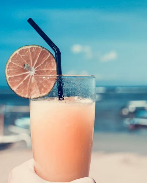 A hand holds a glass with a refreshing drink garnished with a lime slice and a straw, against a background of a beach and ocean scenery.